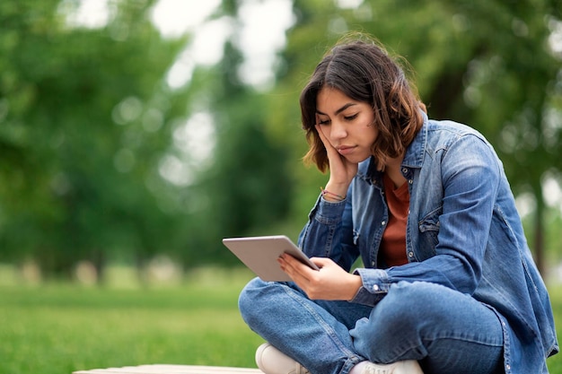 Aluna do Oriente Médio entediada sentada com tablet digital no banco ao ar livre