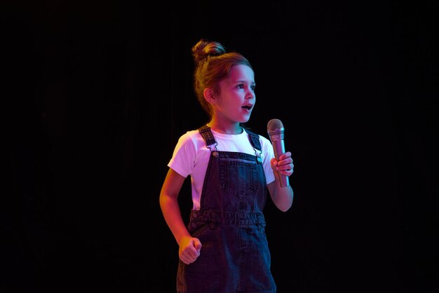 Aluna elegante em camiseta branca e vestido jeans cantando no microfone isolado sobre o escuro