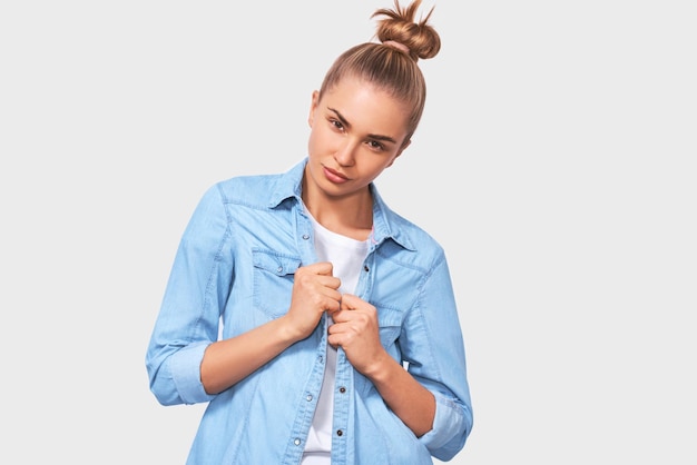 Aluna estudante com penteado de coque olhando seriamente em pé sobre fundo branco do estúdio Jovem mulher vestindo camisa jeans azul posando no fundo do estúdio