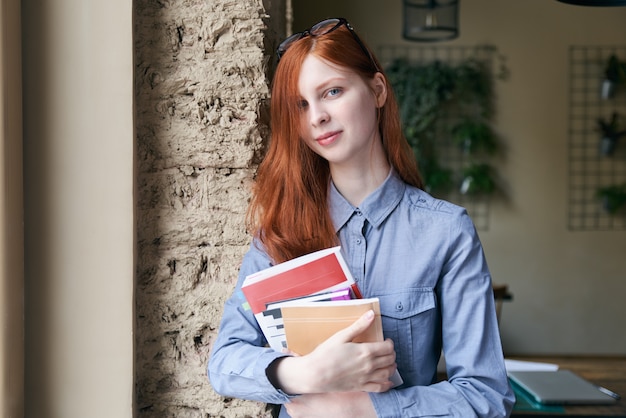 Aluna jovem, com longos cabelos vermelhos posando para um retrato com livros nas mãos com uma expressão confiante relaxada