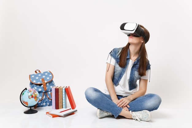 Aluna jovem morena com óculos de realidade virtual, olhando para o lado, apreciando sentar-se perto do globo, mochila, livros escolares isolados
