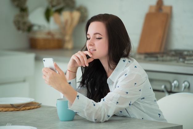 Aluna jovem morena está sentada na cozinha com um smartphone móvel