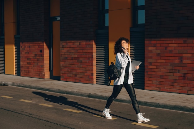 Aluna linda com cabelo encaracolado caminhando enquanto conversa por telefone e segurando um laptop