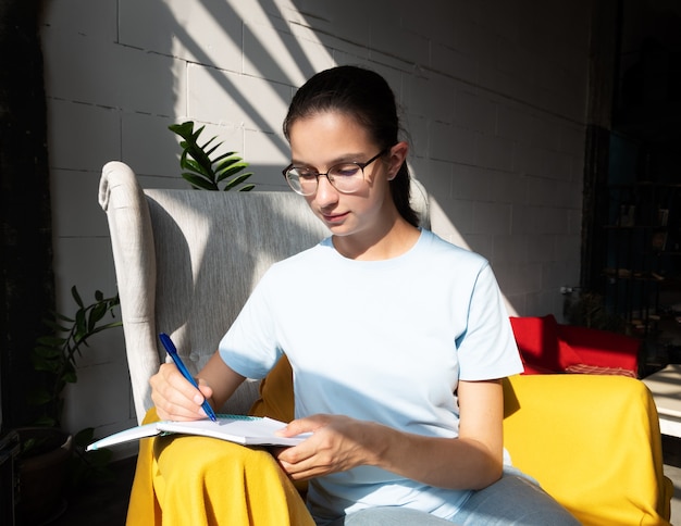 Aluna linda escreve com uma caneta uma tarefa em um caderno enquanto está sentado em uma poltrona em um café com sombras diagonais elegantes, luz forte.