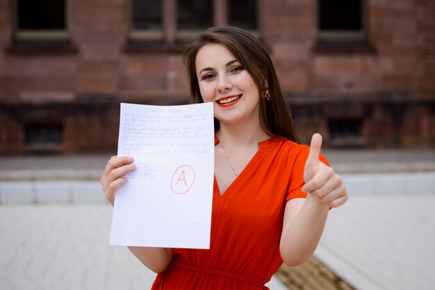 Aluna linda feliz no vestido vermelho, mostrando o papel com bom resultado do teste