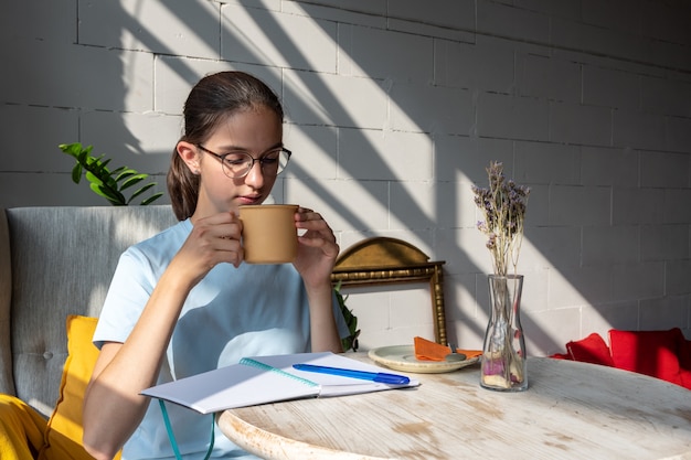 Aluna linda tomando café e escrevendo uma tarefa em um caderno enquanto está sentado em um café com elegantes sombras diagonais, luz forte.