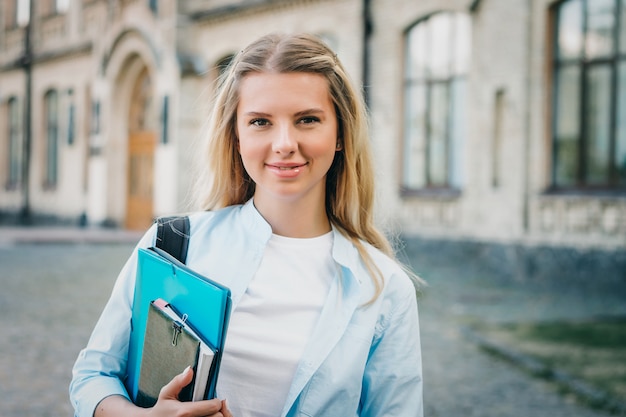 Aluna loira está sorrindo e segurando uma pasta e um caderno nas mãos na universidade