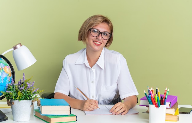 Aluna loira sorridente de óculos, sentada na mesa com as ferramentas da escola, olhando para a câmera, segurando um lápis isolado na parede verde oliva