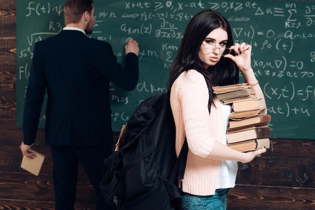 Aluna morena sexy segurando os óculos enquanto carregava uma pilha de livros Jovem universitária no casaco de lã rosa andando na frente da placa verde