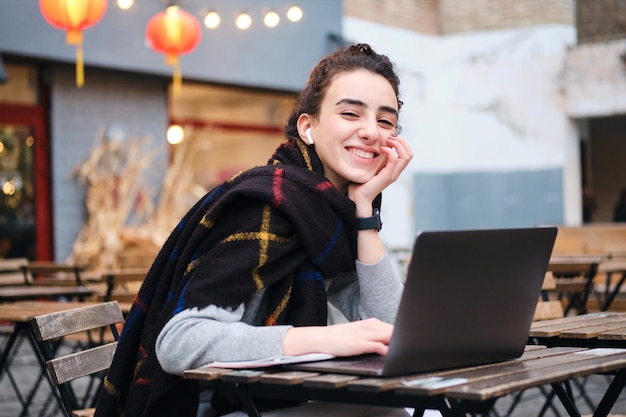 Aluna muito alegre com cachecol estudando alegremente no laptop no café na rua da cidade