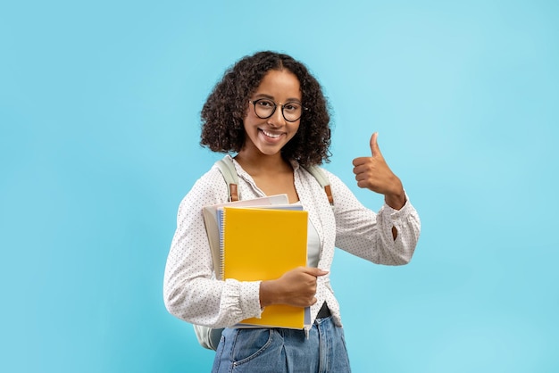 Aluna negra alegre com mochila e notebooks mostrando o polegar para cima recomendando moderno