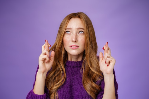 Aluna ruiva fofa e preocupada com sardas estreitando os olhos e focando em fazer um desejo enquanto cruza os dedos para dar boa sorte olhando para o canto superior esquerdo com fé e expectativa sobre a parede roxa.