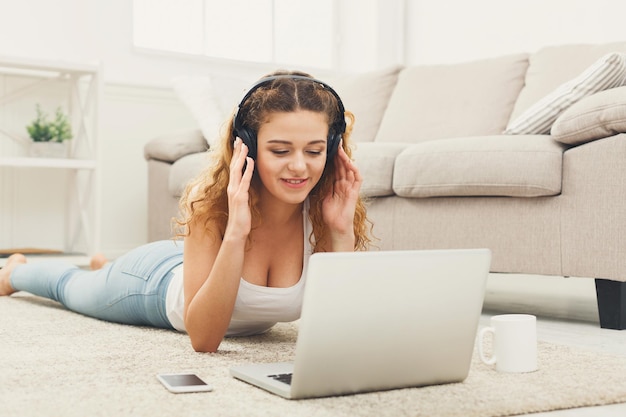 Aluna ruiva sorridente usando laptop para estudar ou trabalhar, deitada no carpete, ouvindo música, copie o espaço