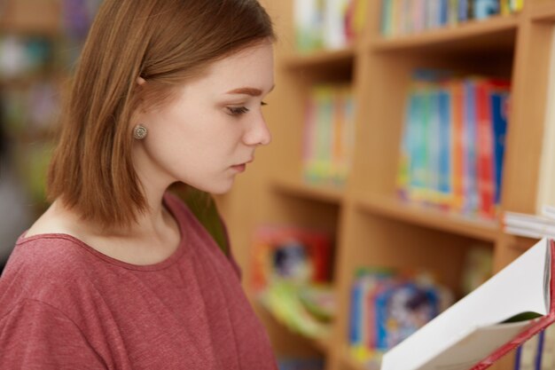 aluna séria inteligente olha atentamente para o livro, fica na biblioteca da universidade