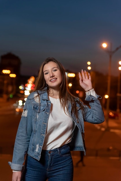 Aluna sorridente caminha na cidade à noite Jovem morena em roupas jeans no fundo da rua da cidade