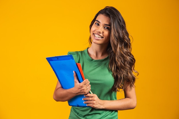 Aluna sorridente com livros escolares nas mãos sobre fundo amarelo.