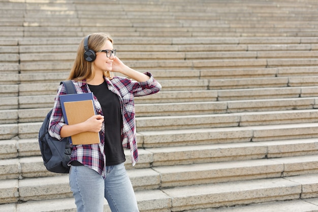 Aluna sorridente com livros sobre fundo de escadas da universidade ao ar livre, olhando para longe, ouvindo música em fones de ouvido durante as férias, descansando no campus. Conceito de educação, espaço de cópia