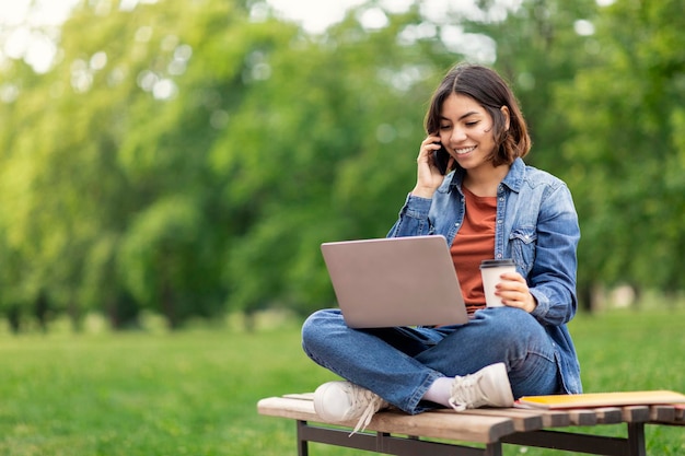 Aluna sorridente do Oriente Médio falando no celular e usando laptop ao ar livre