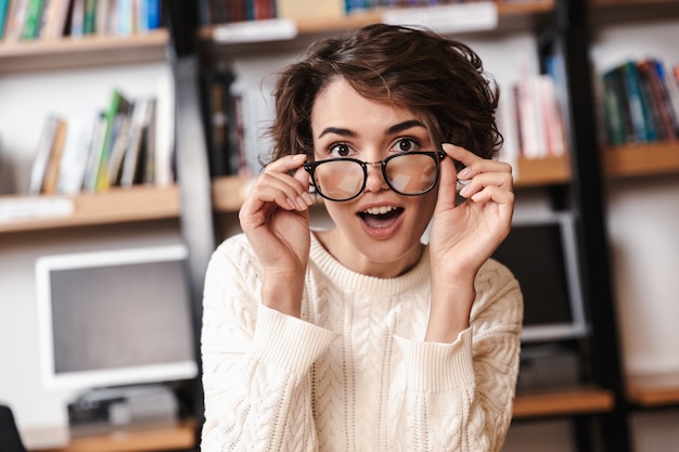 Aluna sorridente, estudando sentado na mesa da biblioteca, usando óculos