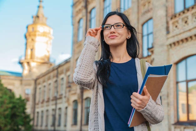 Aluna universitária em óculos sorrindo com livro e bolsa no campus ao ar livre