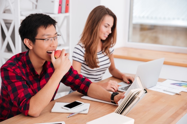 Aluno asiático comendo maçã e lendo enquanto um colega de grupo trabalha no laptop