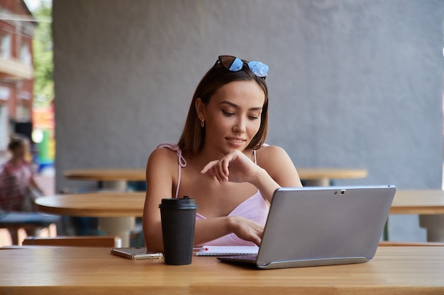 Aluno asiático sentado à mesa do café, estudando com o laptop, fazendo a lição de casa