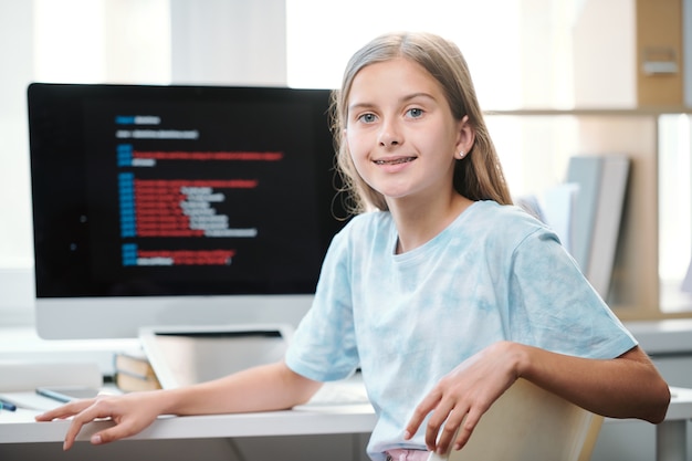 Aluno atraente do ensino médio em roupas casuais olhando para você enquanto está sentado na mesa em frente ao monitor do computador