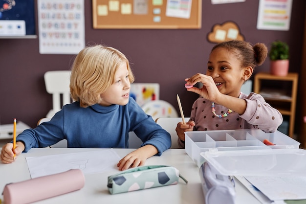 Aluno bonito da escola primária usando apontador de lápis enquanto está sentado na mesa ao lado do adorável scho loiro