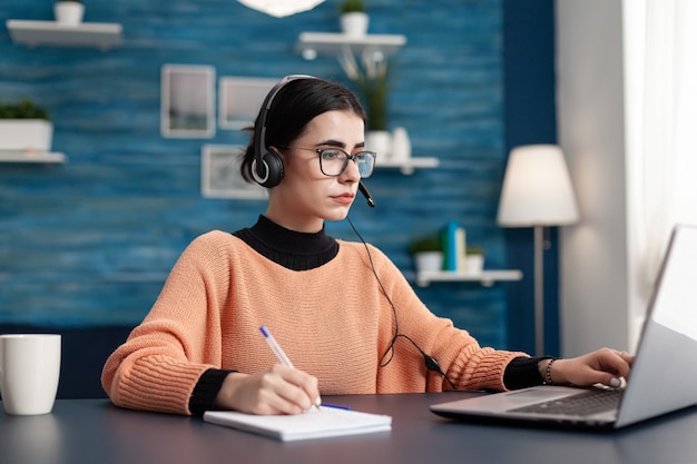 Aluno com fones de ouvido escrevendo notas no caderno enquanto pesquisa informações de comunicação usando o computador laptop. adolescente concentrado fazendo lição de casa enquanto está sentado à mesa na sala de estar