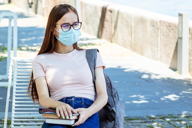 Aluno com uma máscara com livros e uma mochila em um banco do parque no verão proteção contra adolescentes e crianças cobiçosos