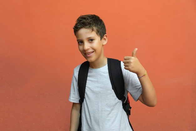 Aluno de escola primária com o polegar para cima isolado contra uma parede laranja de volta ao conceito da escola
