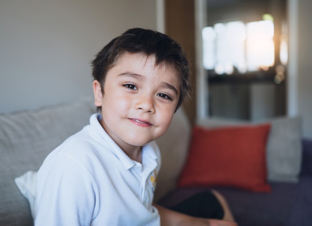 Aluno de rosto closeup olhando para a câmera com uma cara sorridente, Criança feliz relaxando em casa depois de voltar da escola, Criança positiva de retrato interno sentado no sofá na sala de estar