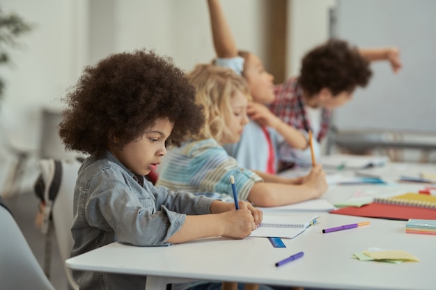 Foto aluno diligente escrevendo em seu caderno enquanto estudava sentado à mesa no ensino fundamental