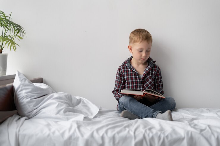 Foto aluno em casa em quarentena. ensino à distância. um menino loiro está sentado na cama lendo um livro. a criança fica em casa e faz o dever de casa sozinha. descanse da escola antes das férias.