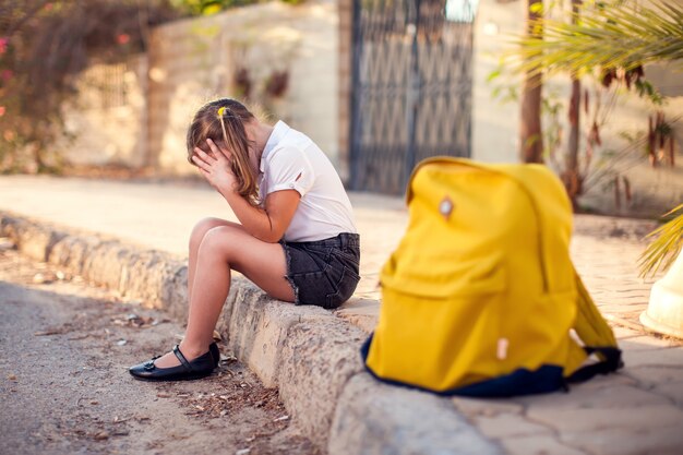 Aluno infeliz com mochila sentado ao ar livre. menina sentindo estresse. conceito de crianças e emoções