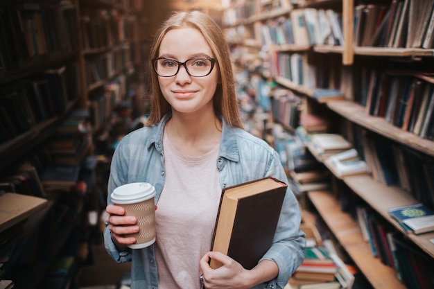 Aluno inteligente e inteligente está de pé na biblioteca pública. Ela está segurando uma xícara de café e um livro ao mesmo tempo. Menina está olhando para a frente com um sorriso no rosto.