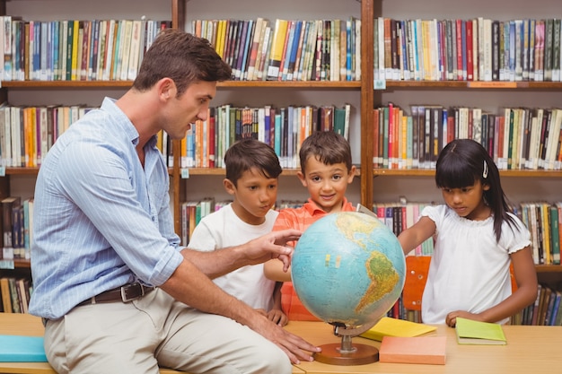 Alunos bonitos e professor que olham o globo na biblioteca