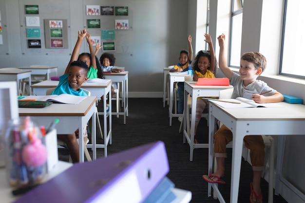 Alunos de escolas primárias multirraciais sorridente com as mãos levantadas sentados na mesa na sala de aula