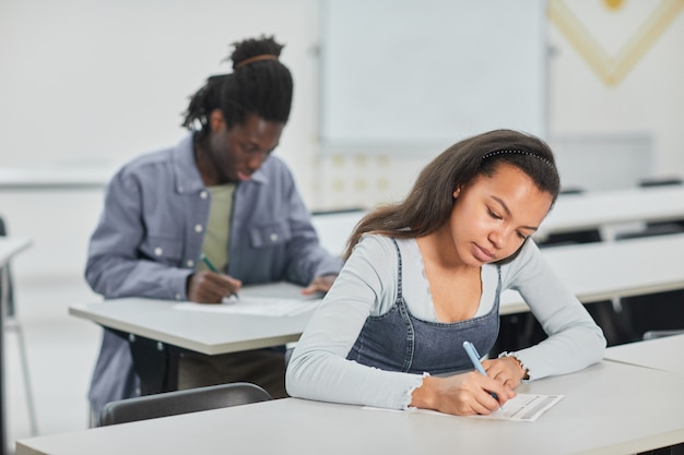 Alunos fazendo teste em sala de aula com foco em uma jovem afro-americana sentada na mesa em frente, copie o espaço