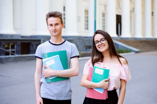 Alunos masculinos e femininos posando perto de universidade convencional com materiais de aprendizagem