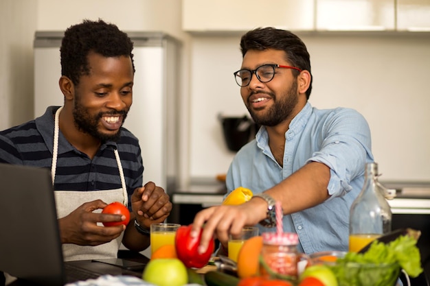 Alunos multiétnicos alegres, cozinhando e se divertindo na cozinha. Cara afro-americano e seu amigo indiano sentado à mesa assistindo a um programa de TV online no laptop e rindo.