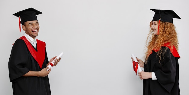 Alunos pretos e ruivos em vestidos de formatura e bonés quadrados ficam em pé, frente a frente, segurando seu querido diploma. Banner com espaço de cópia