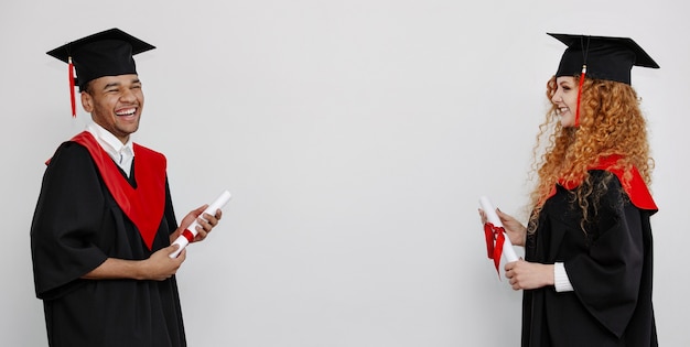 Alunos pretos e ruivos em vestidos de formatura e bonés quadrados ficam em pé, frente a frente, segurando seu querido diploma. banner com espaço de cópia