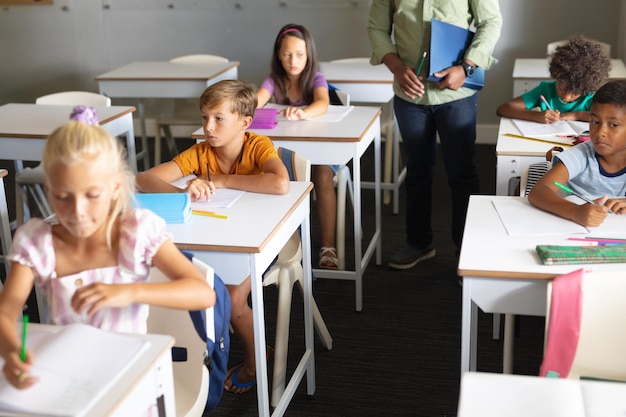 Alunos primários multirraciais estudando em uma mesa com um jovem professor afro-americano na aula. inalterado, educação, aprendizagem, infância, ocupação, ensino e conceito escolar.