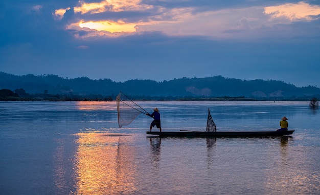 Am frühen Morgen vor Sonnenaufgang wirft ein asiatischer Fischer auf einem Holzboot ein Netz aus, um Süßwasserfische in einem natürlichen Fluss zu fangen.