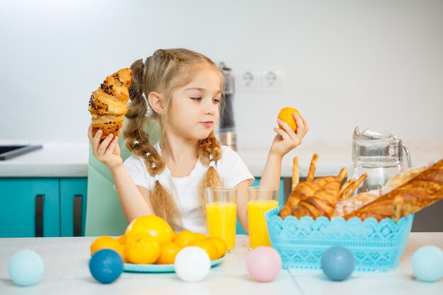 Am Küchentisch sitzt ein kleines Mädchen von sieben Jahren, das ein weißes T-Shirt trägt. Hält Mandarinen-Mandarinen und ein frisch gebackenes Croissant mit Schokolade