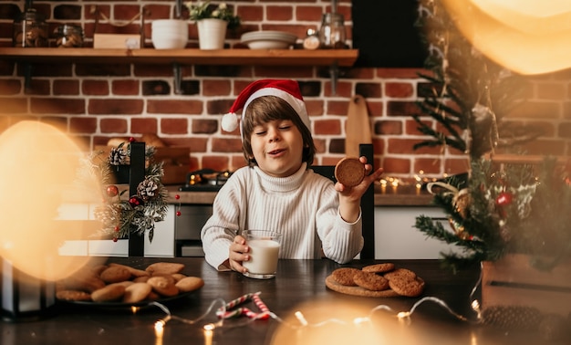 Am Küchentisch sitzt ein Neujahrskind in einem weißen Pullover und einer roten Weihnachtsmütze mit Milch und Keksen