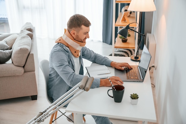 Am Tisch mit Laptop Mann mit Krücken ist zu Hause und hat Verletzungen