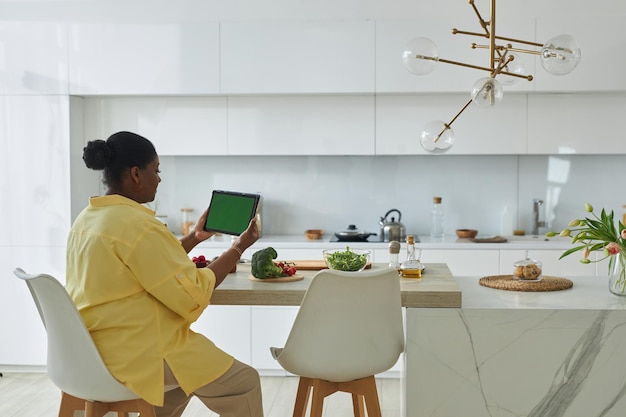Ama de casa afroamericana leyendo receta en tablet pc mientras prepara comida en la cocina