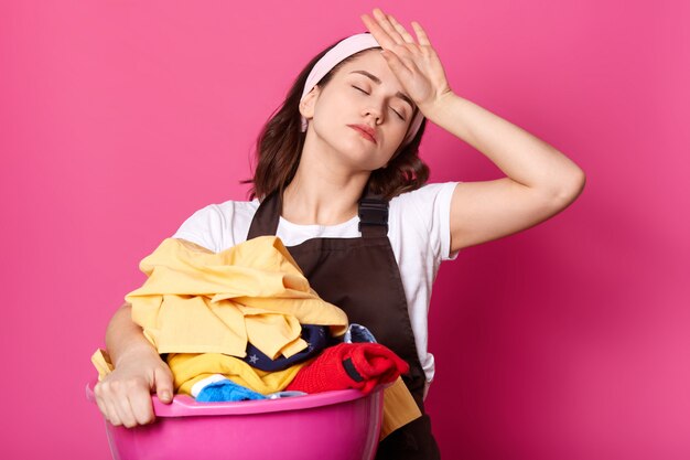 Ama de casa de aspecto agradable con aspecto molesto y cansado, usa un delantal marrón y una camiseta blanca informal, posa contra la pared rosa, se siente agotada después del trabajo duro en el día en casa. Copia espacio